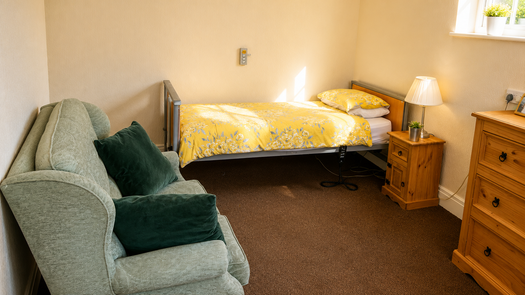 A bright, well-furnished resident's room with yellow floral bedding, a green armchair, and pine furniture.