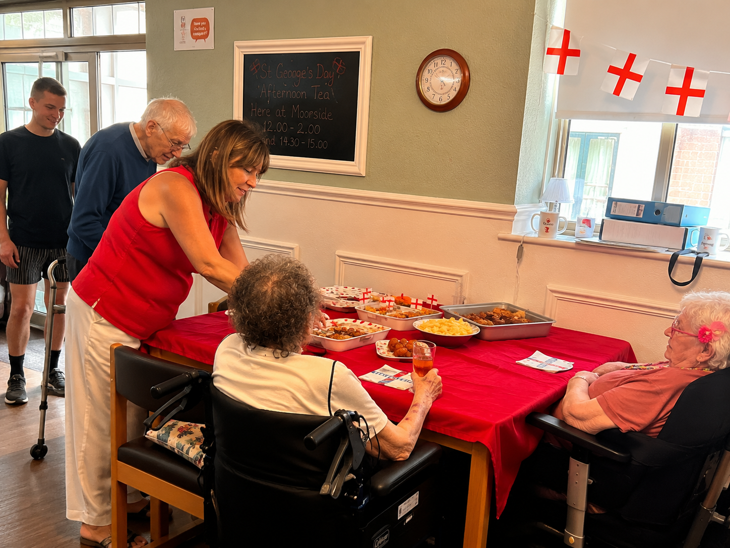 A staff member in red serves residents at the St George's Day Afternoon Tea, with trays of party food on a red-draped table.